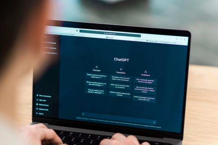 Foto em close de uma pessoa utilizando um notebook que exibe a interface inicial do ChatGPT, mostrando colunas de exemplos, capacidades e limita&ccedil;&otilde;es.