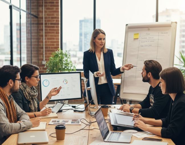 Mulher executiva em pé, apontando para um flip chart durante uma reunião de equipe multidisciplinar de agência digital, com cinco colegas sentados à mesa, observando e interagindo com dados em um monitor e notebooks.